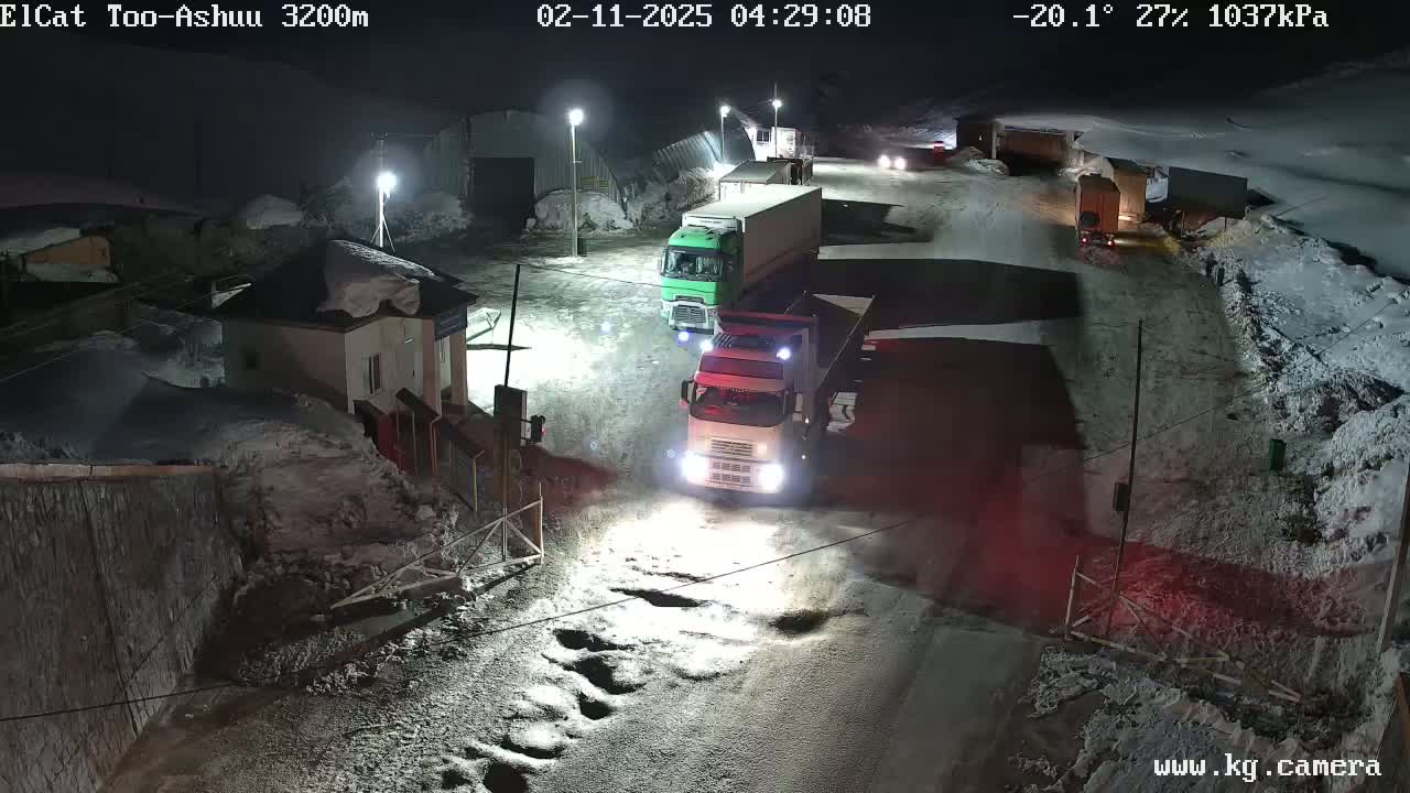 At night, in snowy conditions, several trucks are parked or driving on a road near a building.