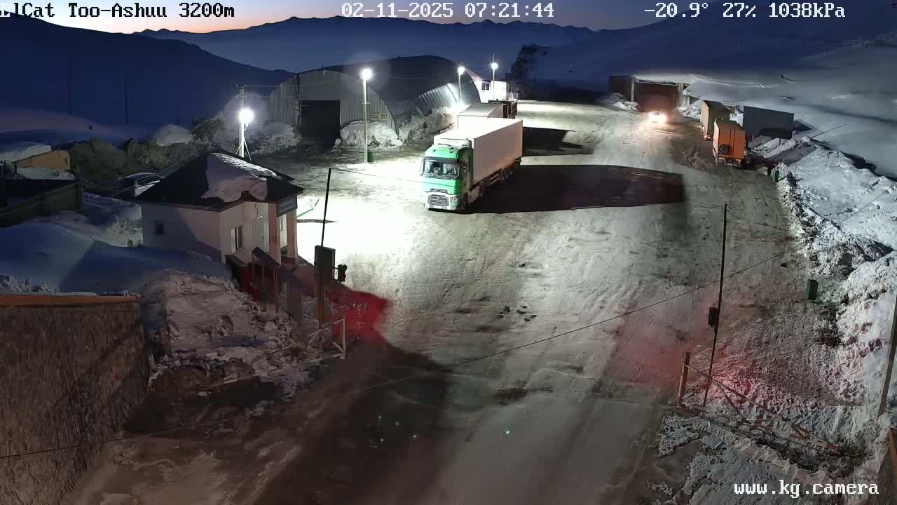 A snow-covered mountain road at night is illuminated by artificial lights, showing several trucks parked near a building.