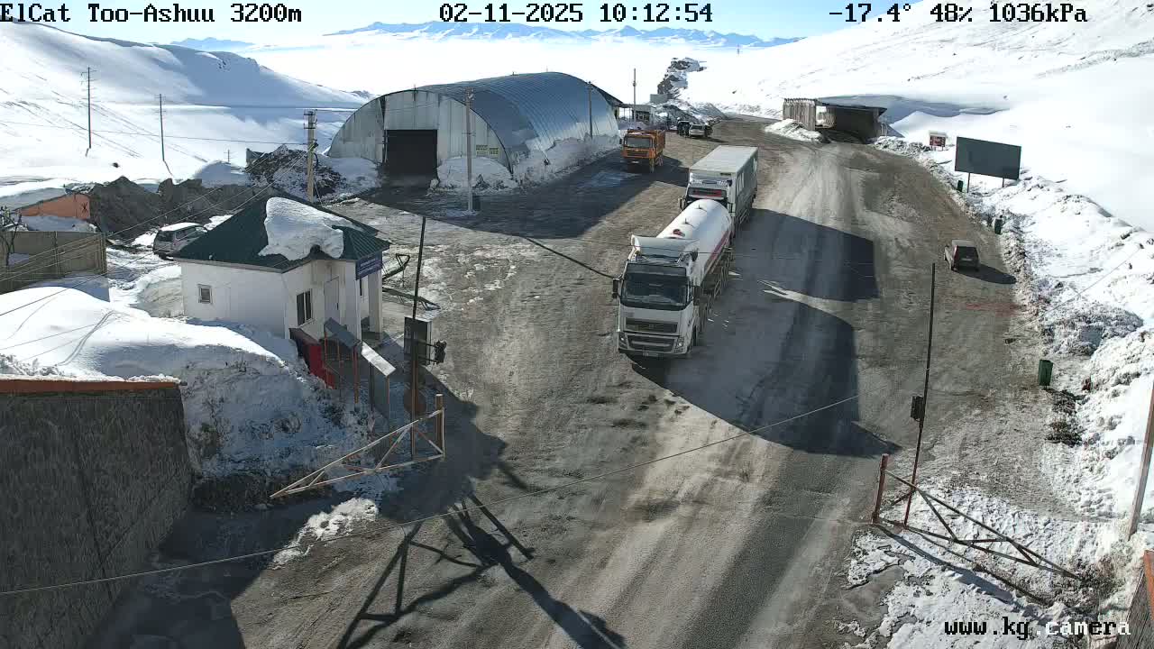A snowy mountain road scene shows several trucks and cars near a small building and a large metal shed under a sunny, cold sky.