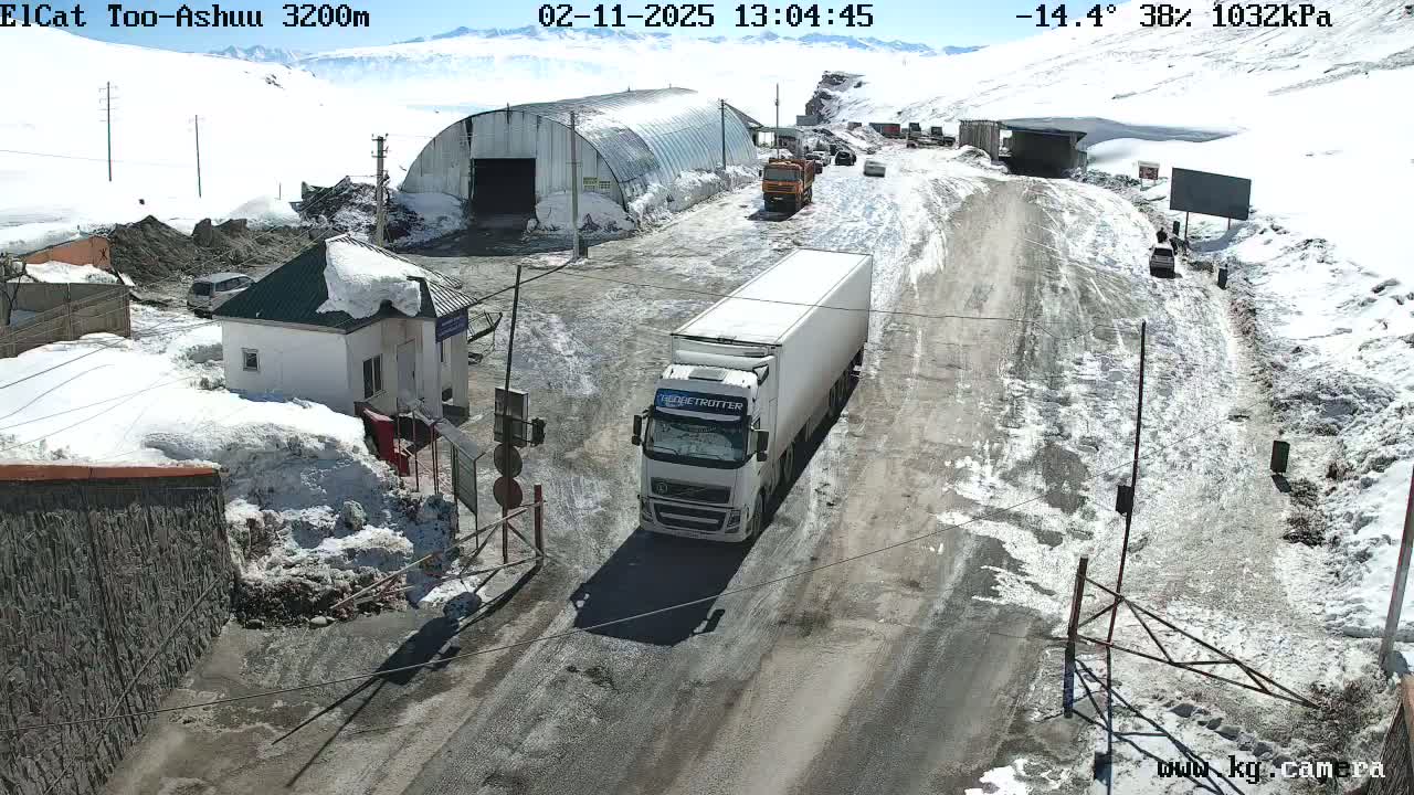 A snowy mountain road with a white semi-truck driving past a border checkpoint building, under partly sunny skies and a temperature of -14.4 degrees Celsius.