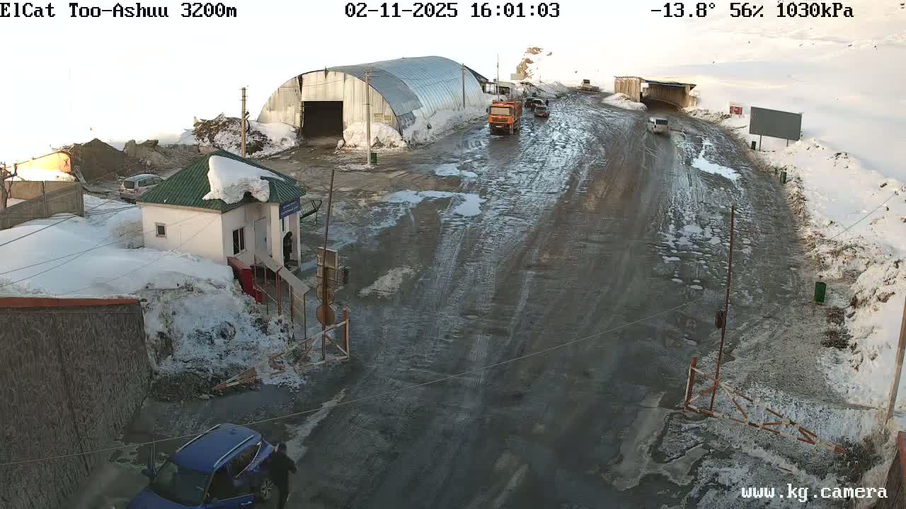 A muddy road in a snowy, mountainous area shows several vehicles near a small building and a large metal structure; the weather is -13.8°C with 56% humidity.