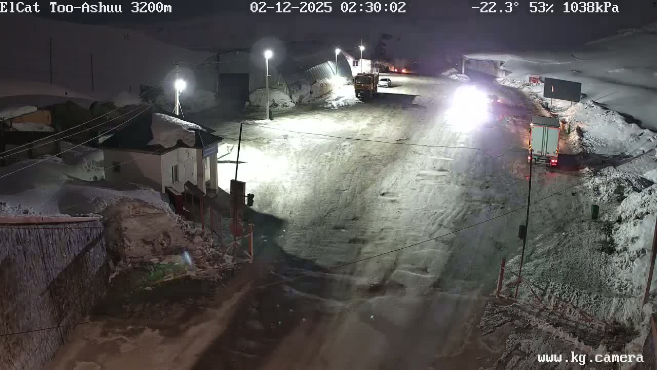 A snow-covered road at night with several vehicles, including trucks, near a building and snow banks under partly cloudy, cold conditions.