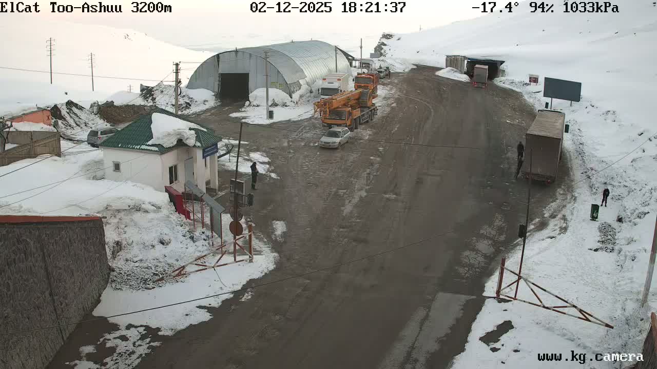 A snowy mountain road intersection with several vehicles, including trucks and a crane, near a small building and a larger metal structure, under a cloudy sky with a temperature of -17.4°C.