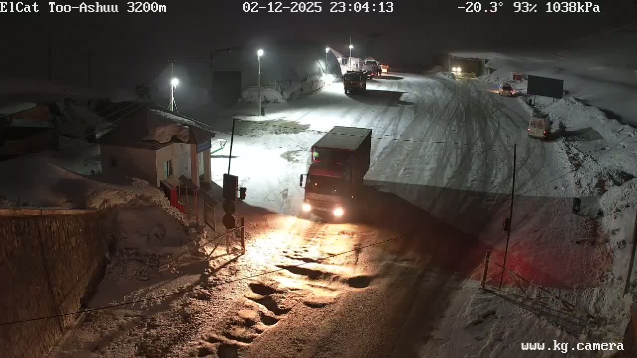 A snow-covered road at night with several vehicles, including trucks, driving on it, near a building.