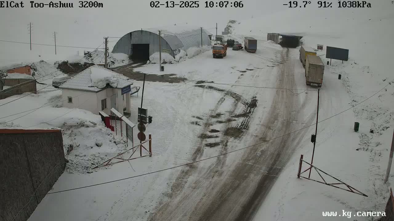A snow-covered mountain pass road with several trucks and a small building, in -19.7°C weather with 91% humidity.