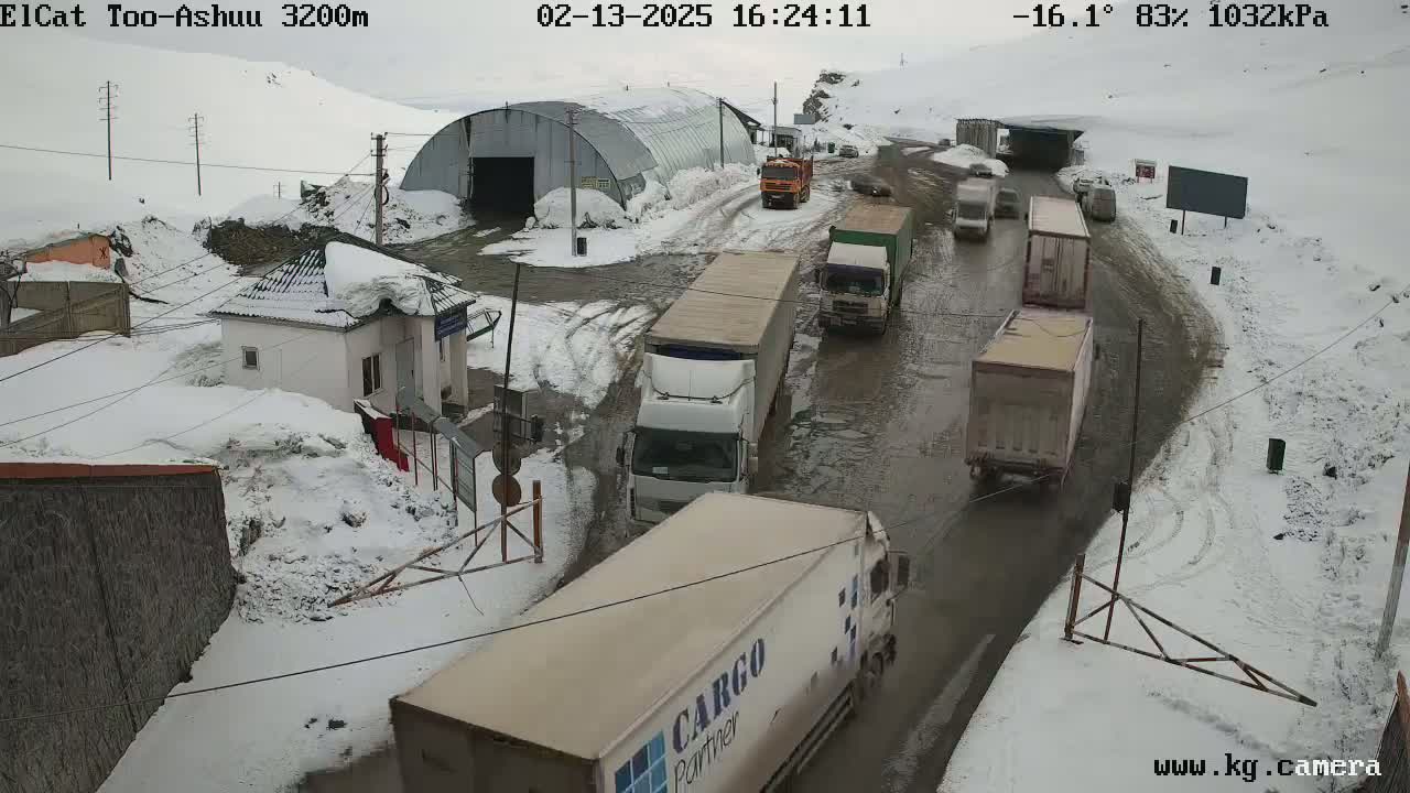 Several trucks navigate a snowy, muddy road near a small building and a large metal structure, under a cloudy sky.
