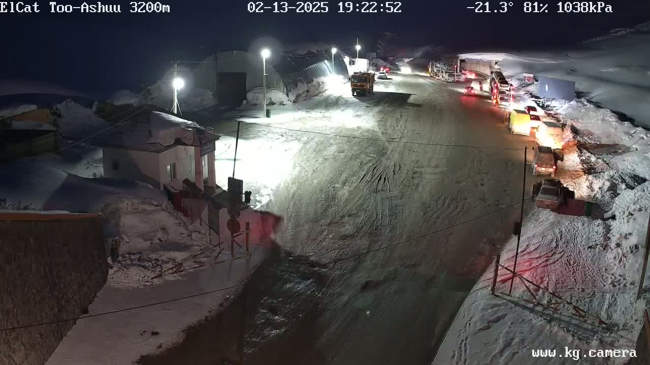 A snow-covered road at night with several vehicles, including trucks and cars, parked and driving along a road near a small building under brightly lit streetlights.