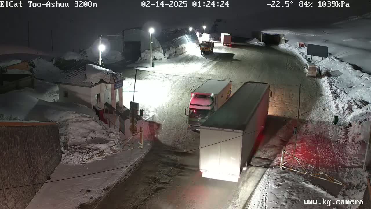 At night, in a snow-covered area with a temperature of -22.5 degrees Celsius, several large trucks and a smaller vehicle are parked near a building.