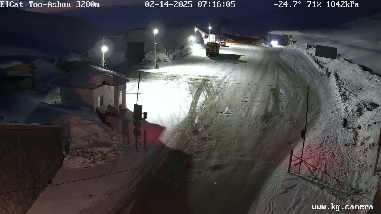 A snow-covered road at night, with a few vehicles and buildings visible under dim lighting.