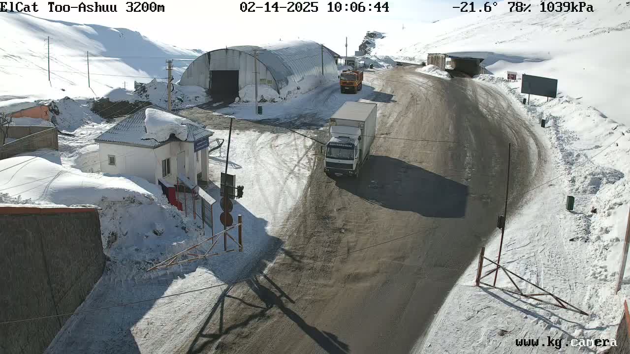 A snowy mountain road with several trucks and a small building, under sunny skies.