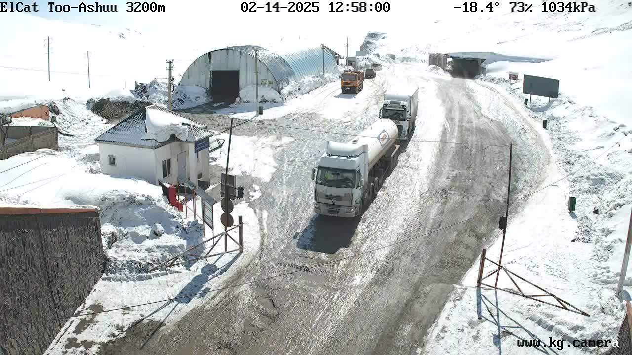 Several trucks are driving on a snow-covered road near a small building and a large metal structure, with a sunny, cold (-18.4°C), and dry (73%) atmosphere.