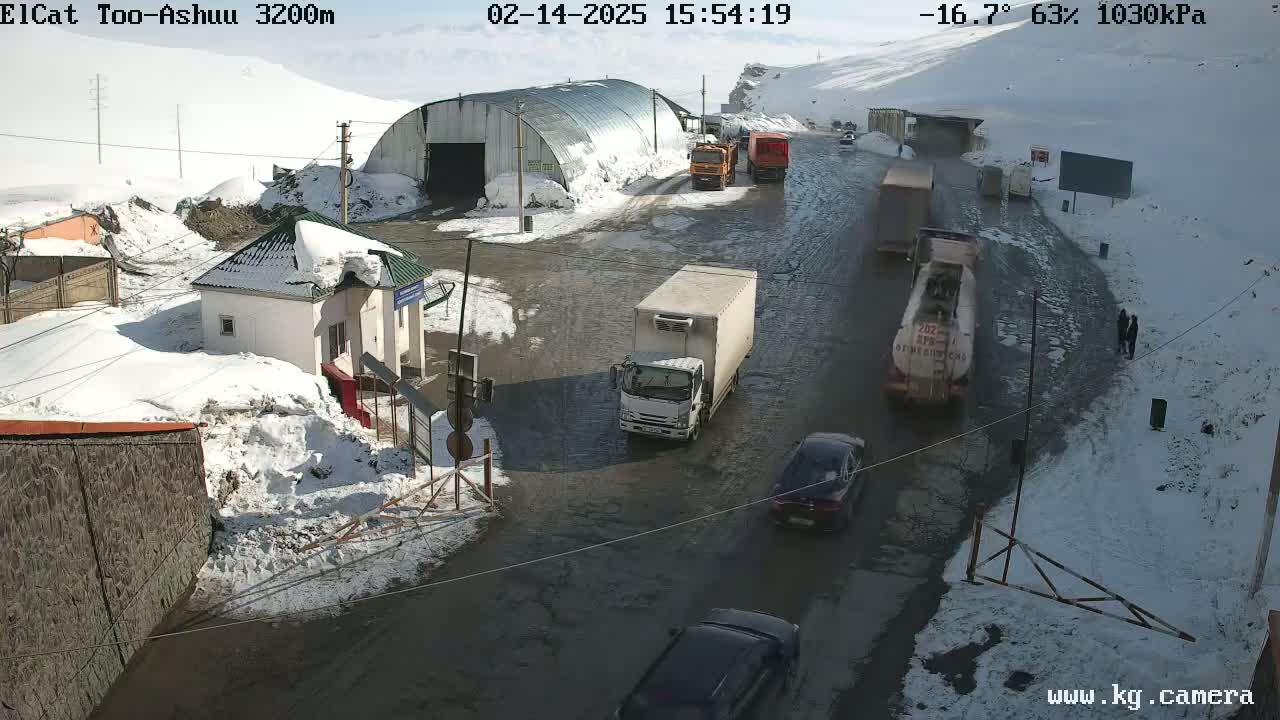A snow-covered mountain pass road with several vehicles, including trucks and cars, navigating a slushy, partially icy roadway under sunny, cold conditions.