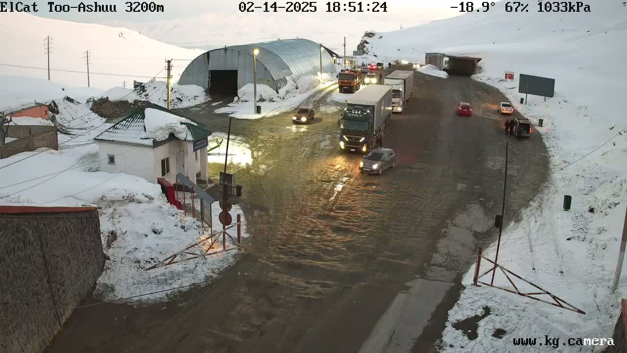 A snowy, wet roadway with several vehicles, including trucks and cars, near a building and a tunnel, under a cloudy sky.
