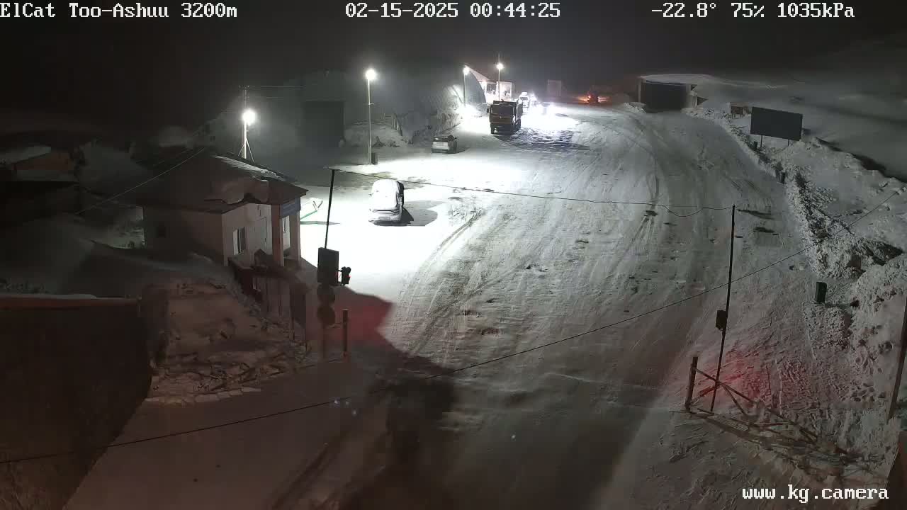 A snow-covered road at night, with a few vehicles visible and buildings in the background under a dark, snowy sky.