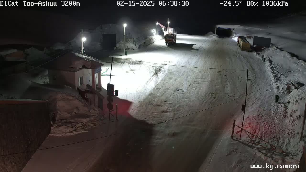 A snow-covered road at night, illuminated by streetlights, with several vehicles parked near a small building and snow banks lining the sides.