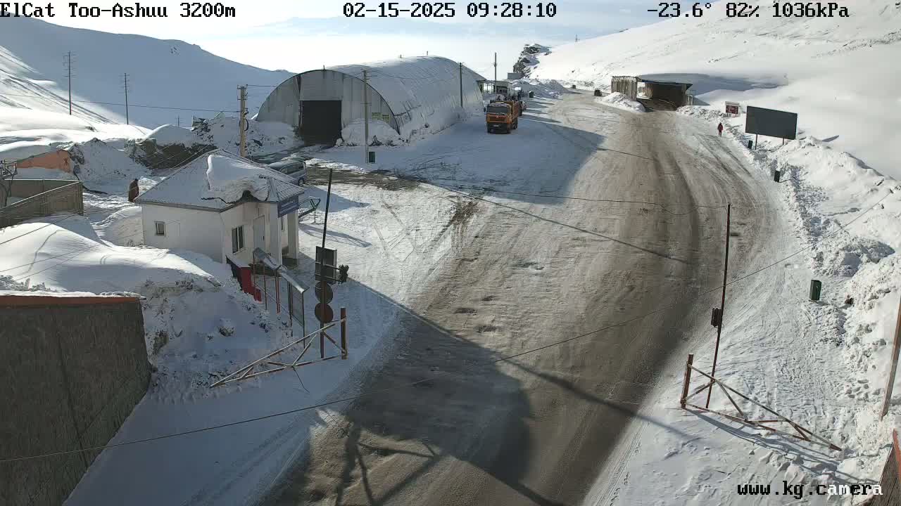 A snowy mountain road scene shows a small building, a large metal structure, and several vehicles, under sunny, cold weather conditions.
