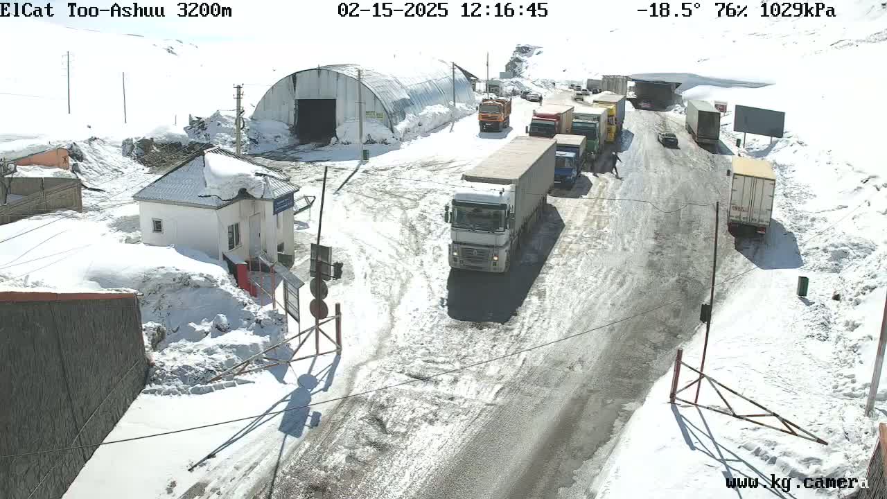 A snowy mountain road with several trucks and cars, a small building, and a large metal structure under a sunny sky.