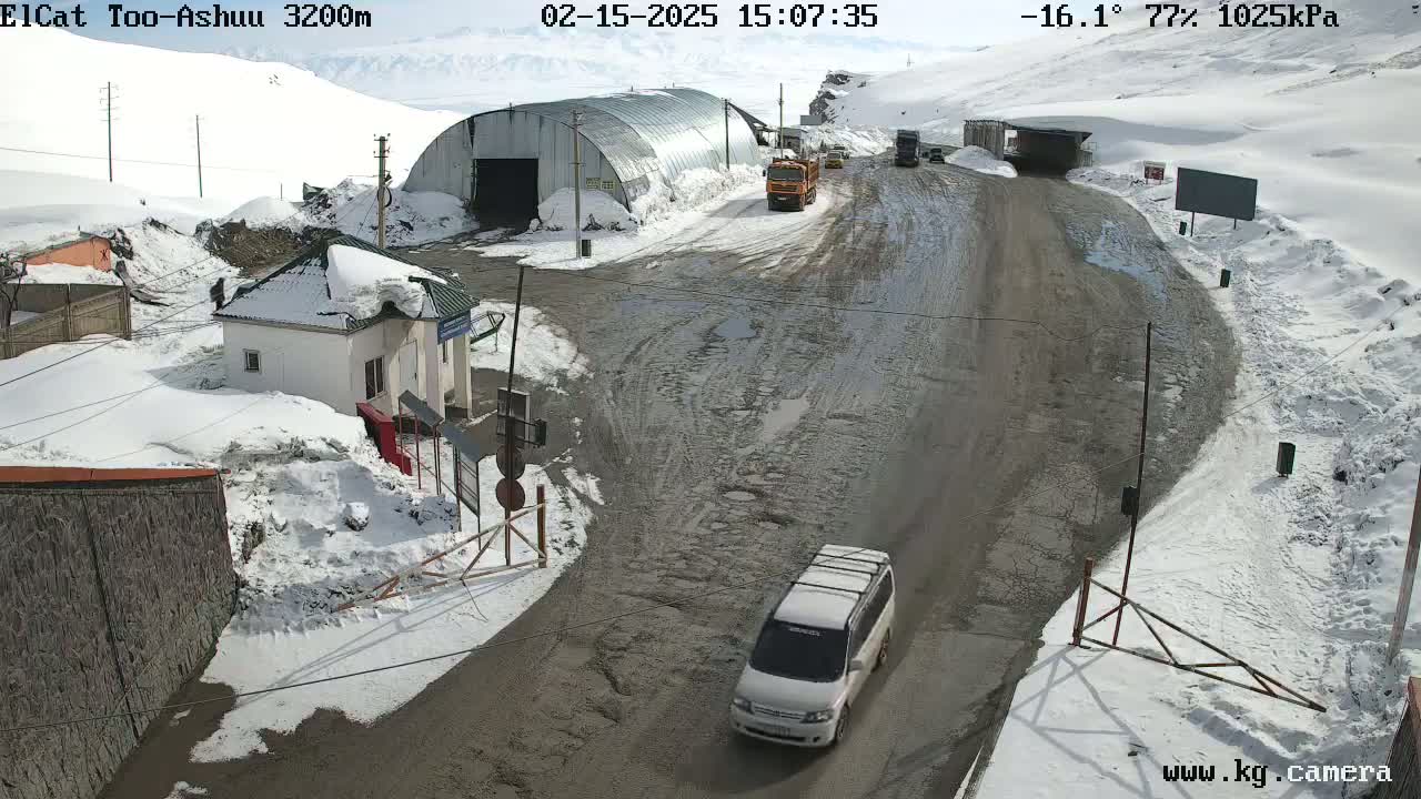 A snow-covered mountain road with several vehicles, a small building, and a large metal structure, under a sunny sky.