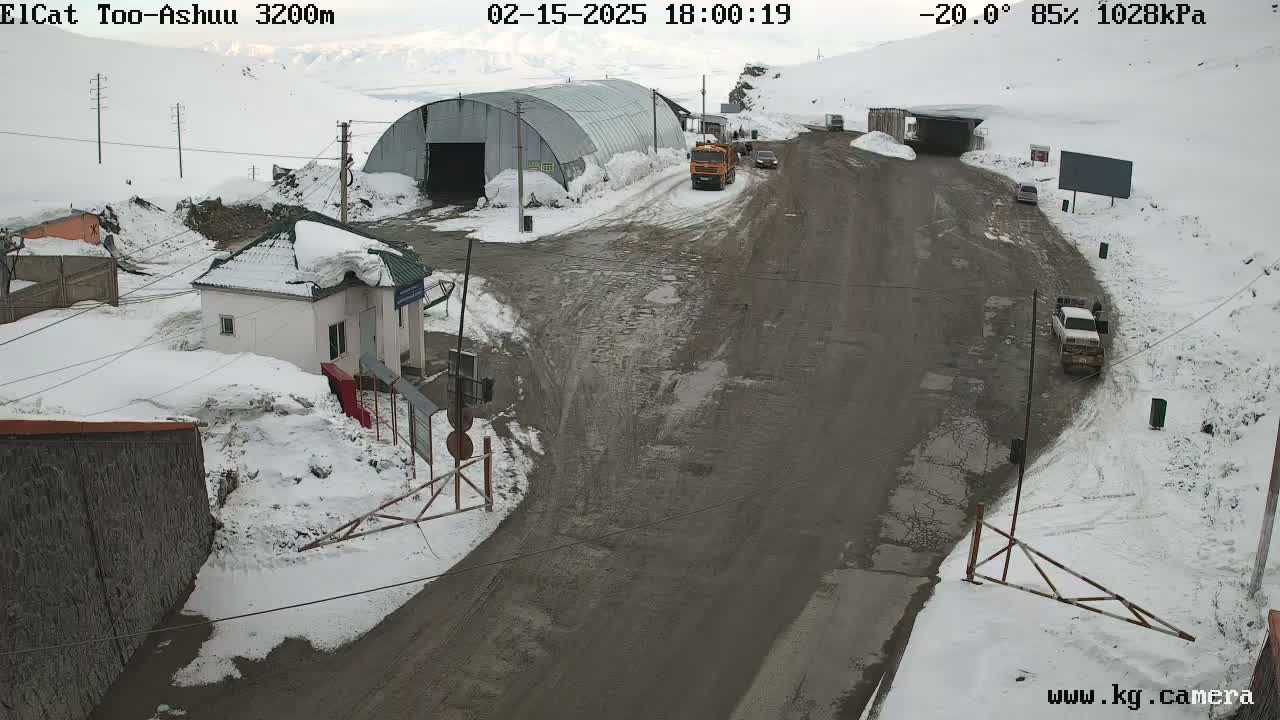 A snow-covered mountain road with several vehicles and buildings, including a small house and a large metal structure, in a -20°C, 85% humidity environment.