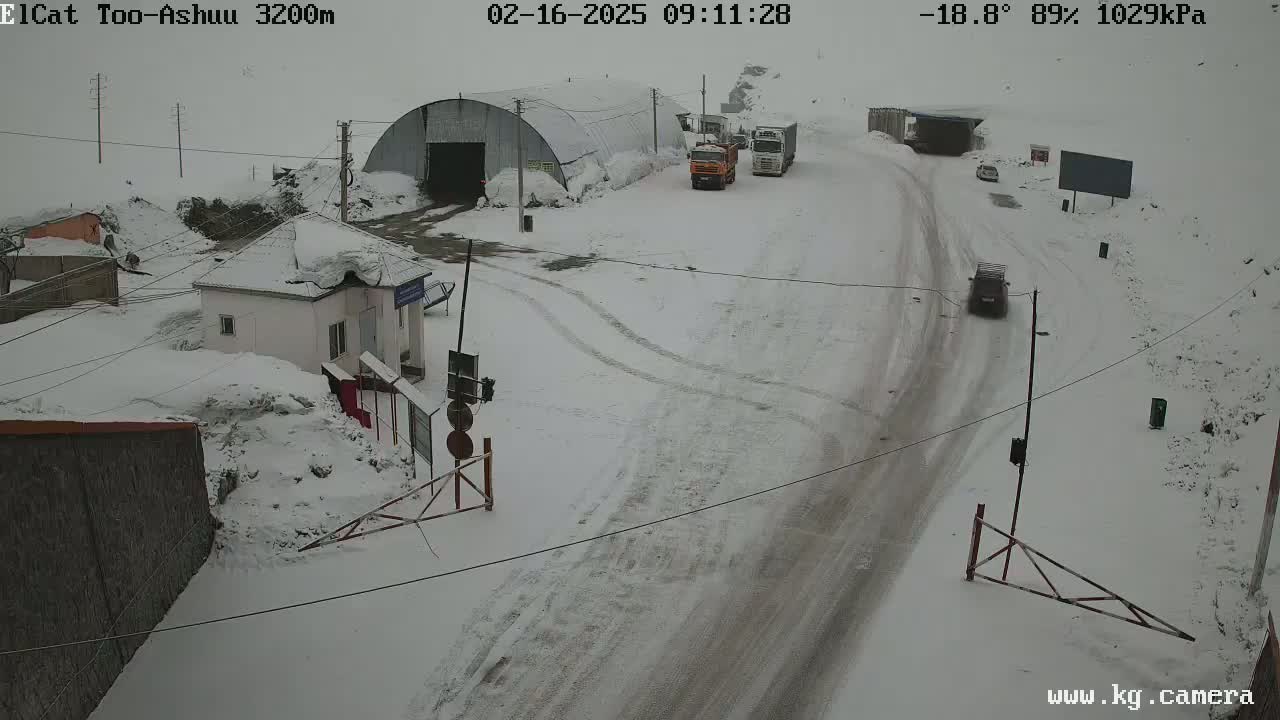 A snow-covered road with several vehicles, a small building, and a large metal structure is visible in a snowy, overcast scene.