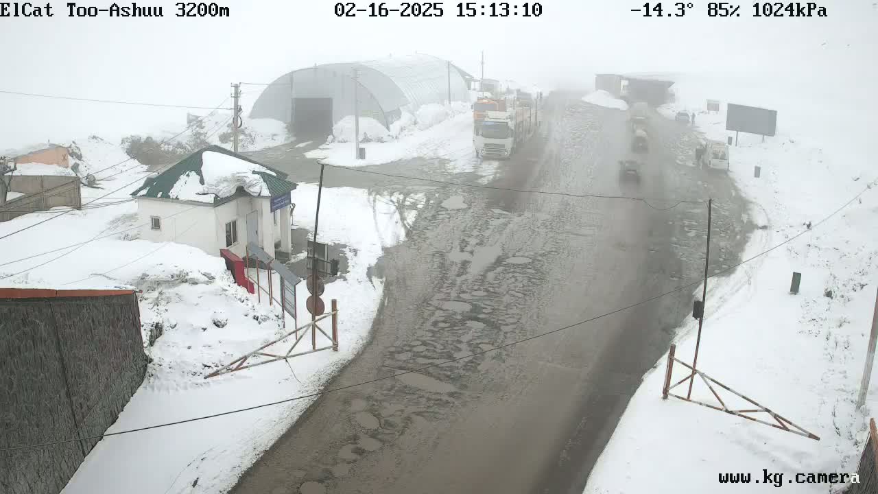 A snowy, foggy road scene shows several vehicles traveling on a road with many potholes, passing by a small building and a large structure; the overall weather is foggy with snow on the ground.