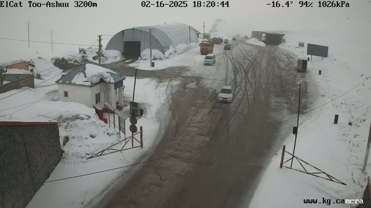 A snow-covered road with several vehicles, a small building, and a large metal structure is visible in a scene of overcast snowy weather.