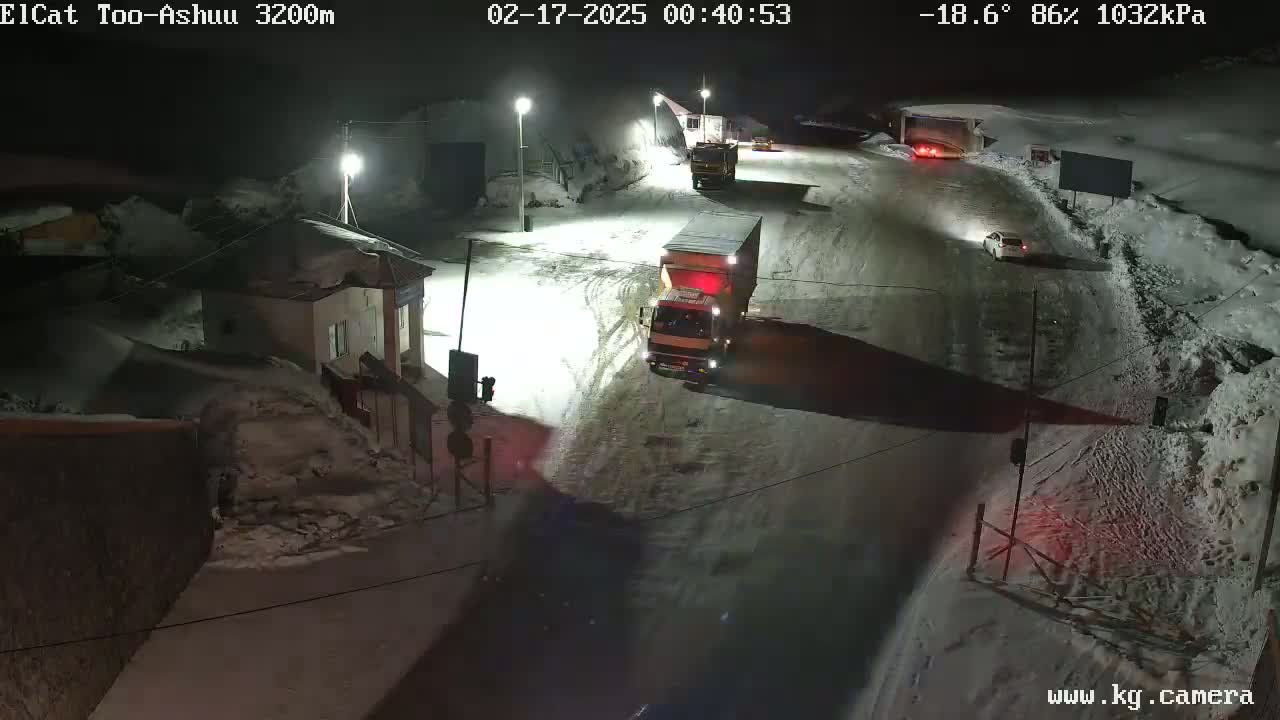 A snow-covered road at night with several vehicles, including trucks and cars, and a small building is visible.