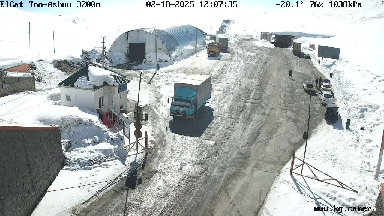 A snow-covered mountain road scene shows several vehicles near a small building, with a sunny sky above.