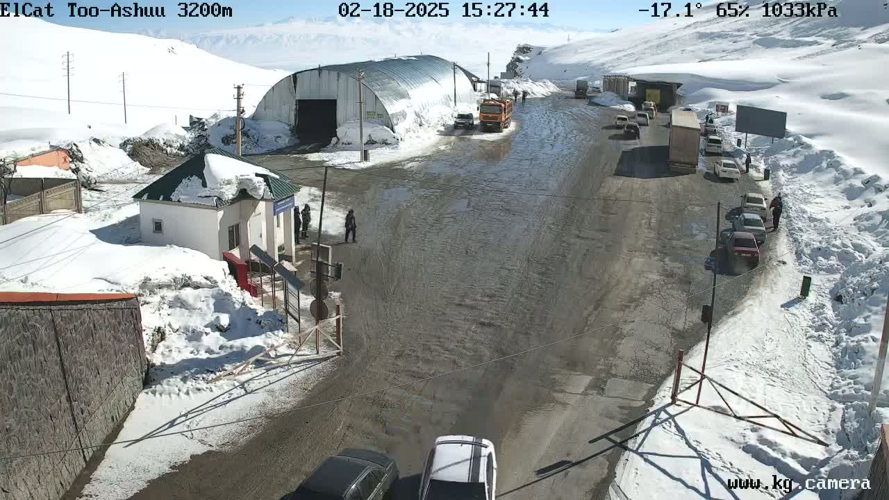 A snowy mountain road with several vehicles and a small building, under a partly sunny sky.