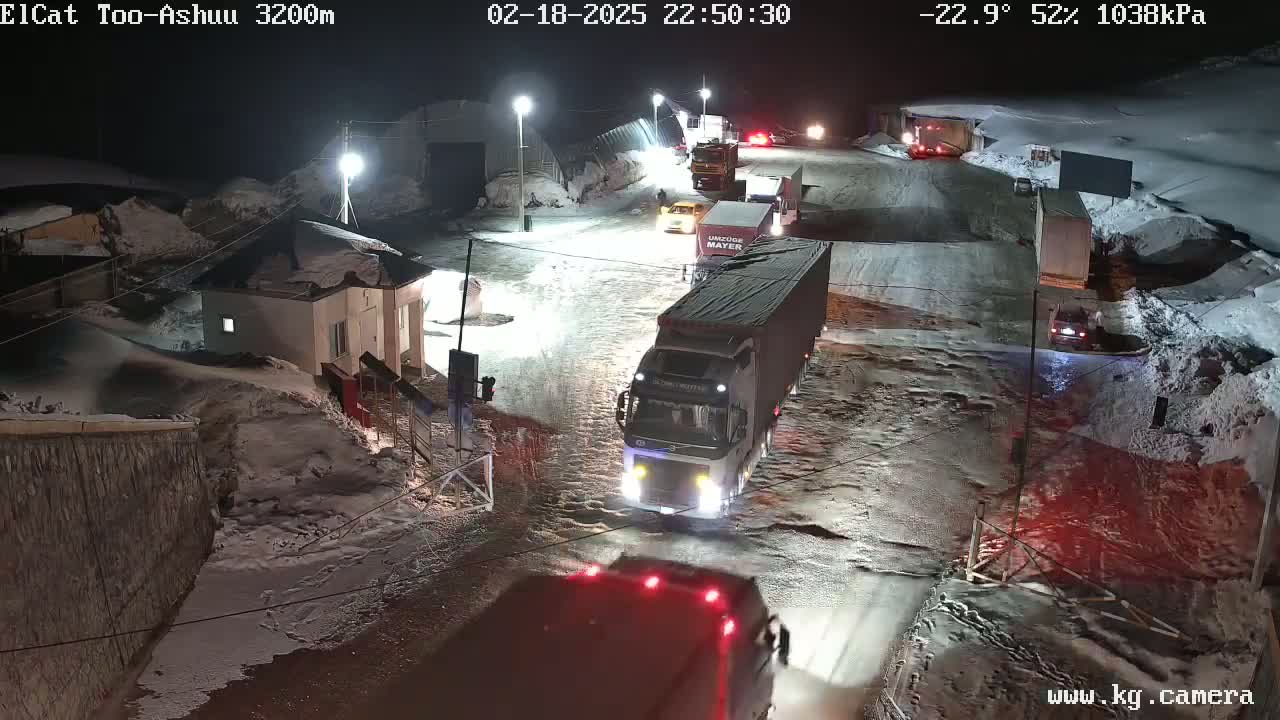 At night, in snowy conditions with a temperature of -22.9 degrees Celsius, several vehicles, including large trucks, are traveling on a road near a small building.