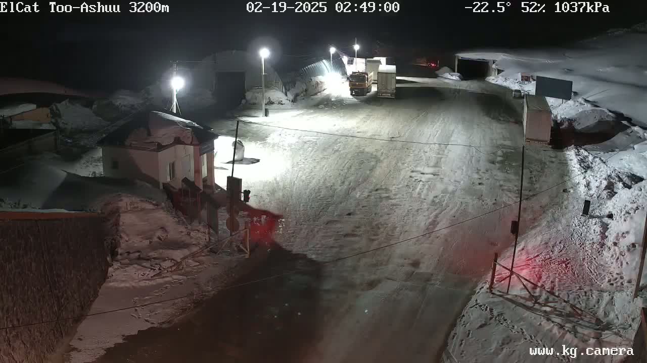 A snow-covered roadway at night is illuminated by several streetlights, with a small building and two trucks visible near a gated area.
