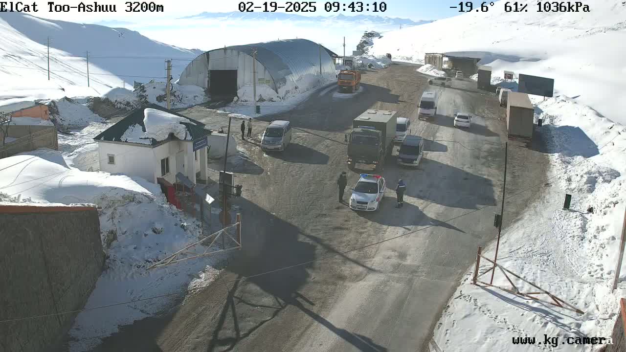A snowy mountain pass road with several vehicles, including a police car, stopped near a small building and a larger structure, under a sunny sky.