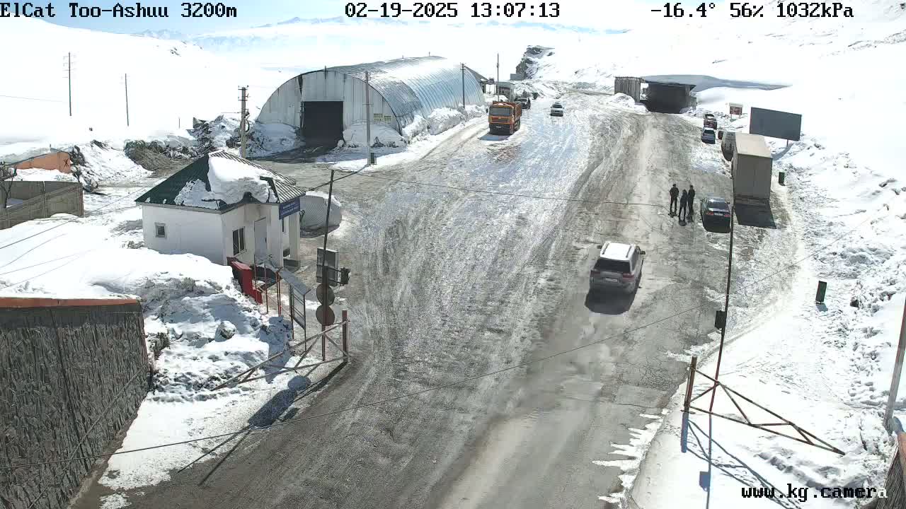 A snow-covered mountain road with several vehicles and a small building, under a partly sunny sky with a temperature of -16.4°C.