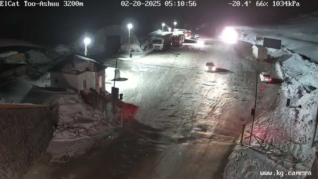 A snow-covered road at night with several vehicles, including trucks and cars, near a small building and under streetlights.