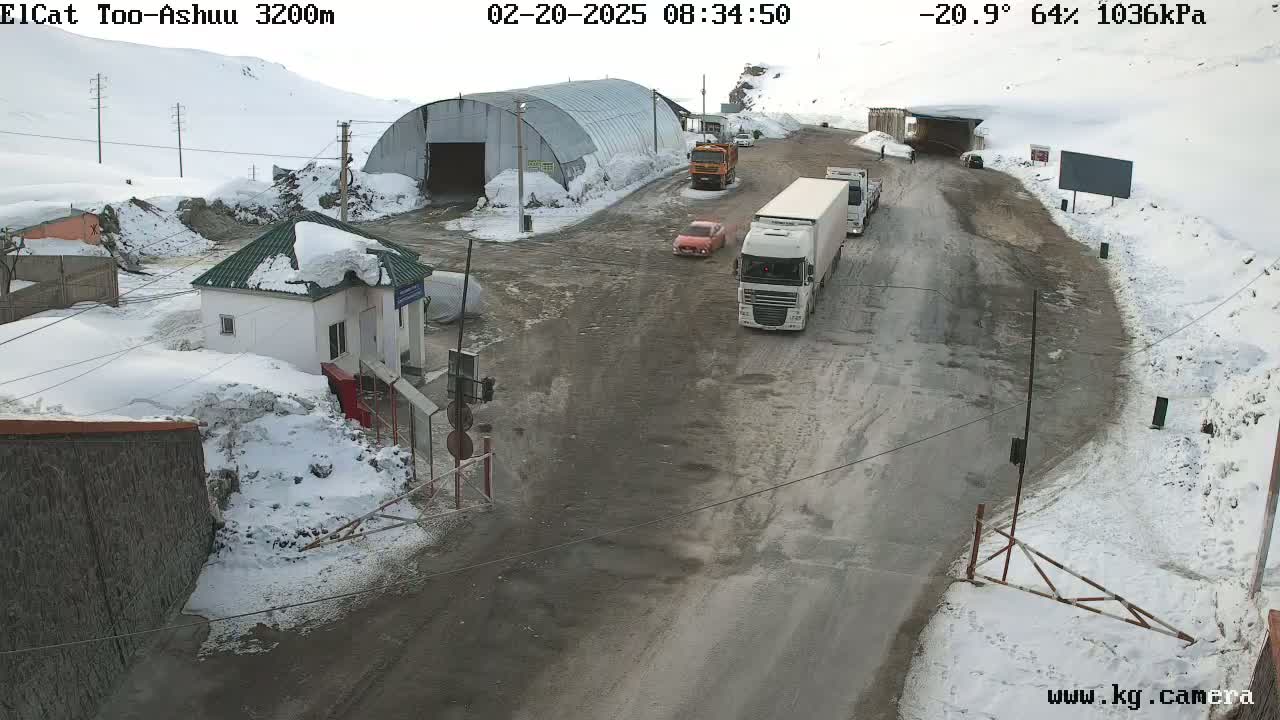 A snowy mountain pass road scene shows several vehicles near a border checkpoint building and a large metal shed, under a partly cloudy sky.