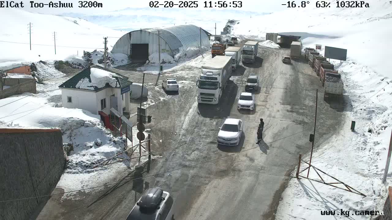 A snowy mountain pass road with several vehicles, including cars and trucks, at a checkpoint building, under a partly sunny sky.