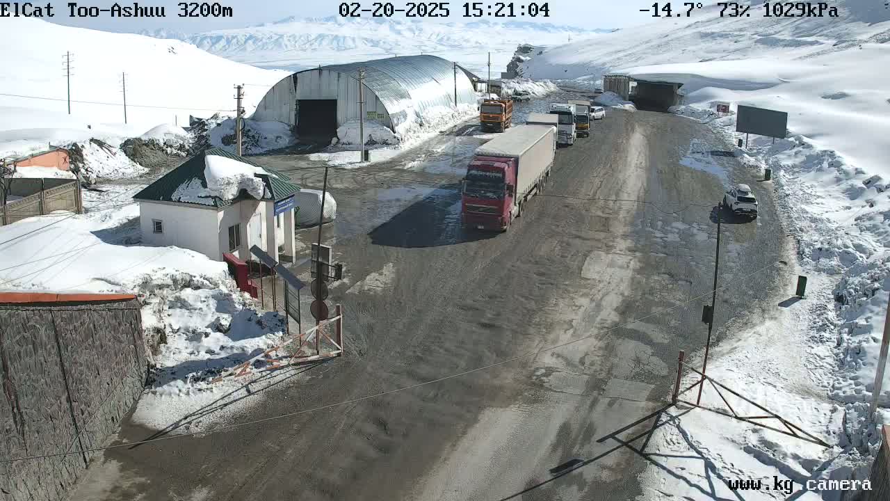A snow-covered mountain pass road with several trucks and cars, a small building, and a large metal structure in the background, under sunny skies.