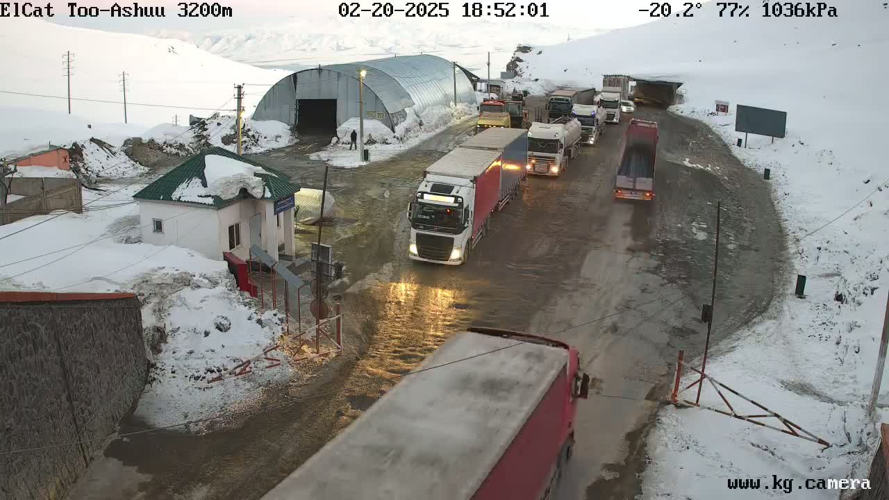 A line of trucks travels on a slushy, snow-covered road past a small building and a large metal structure in a mountainous area.