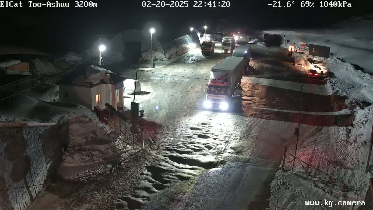 At night, several trucks and cars are stopped on a snow-covered road near a small building, under a mostly clear, cold (-21.6°C) and dry (69% humidity) sky.