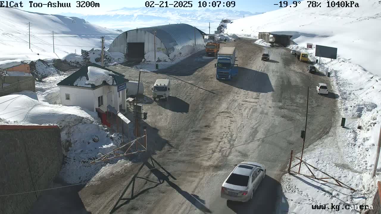 A snow-covered mountain pass road with several vehicles near a checkpoint building and a large metal structure, under a sunny sky.