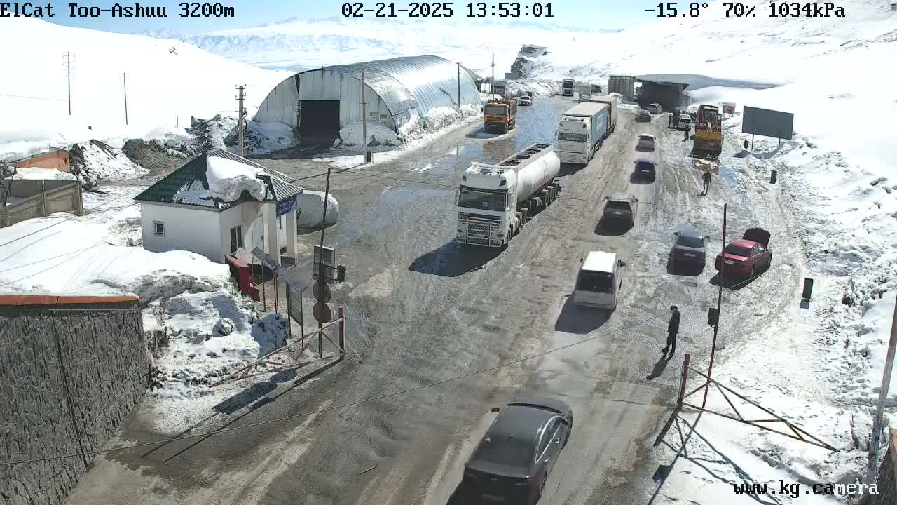 A snowy mountain road with several vehicles, including trucks and cars, passing through a checkpoint near a metal building.