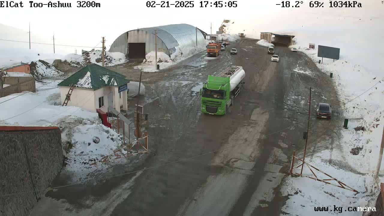 A snowy mountain road with several vehicles, including a green tanker truck, near a small building and a larger metal structure.