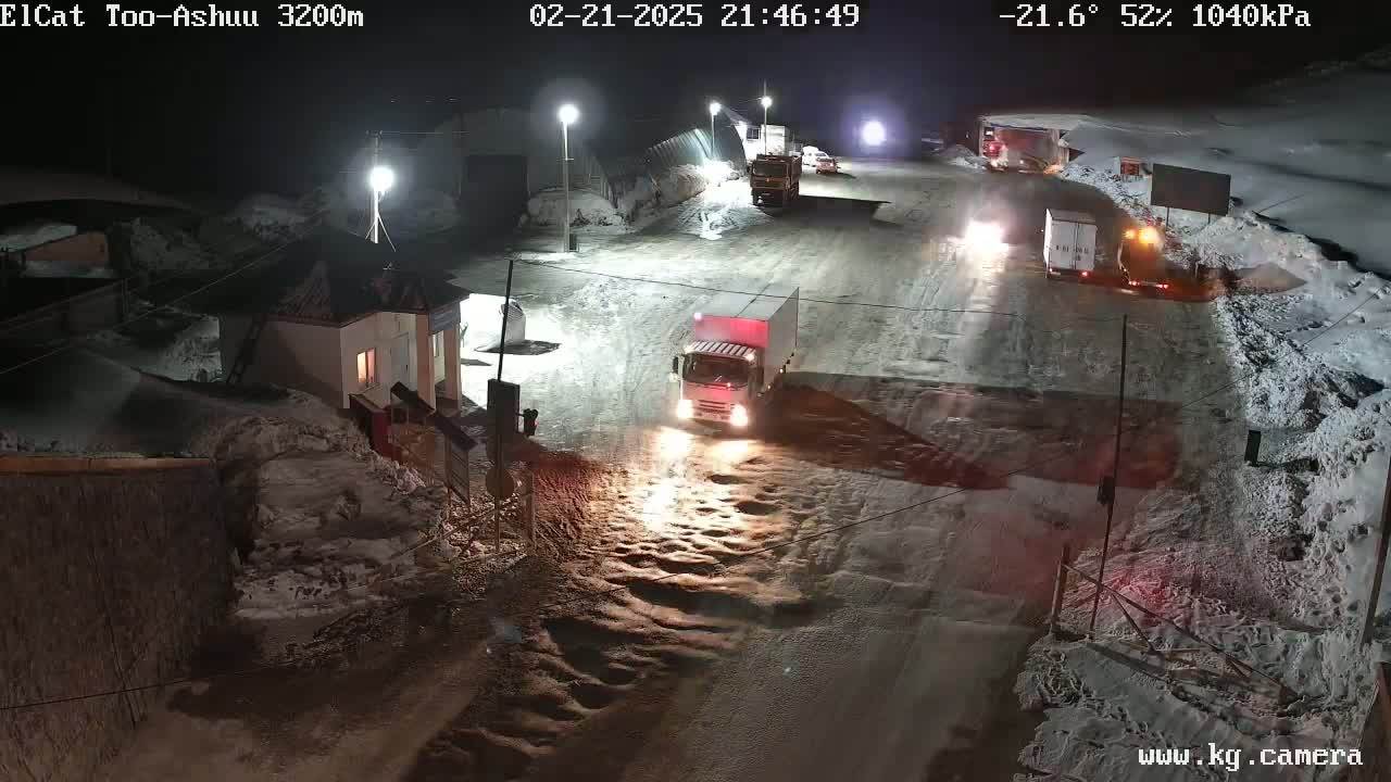 At night, several vehicles are parked and driving on a snow-covered road near a small building, under artificial light.