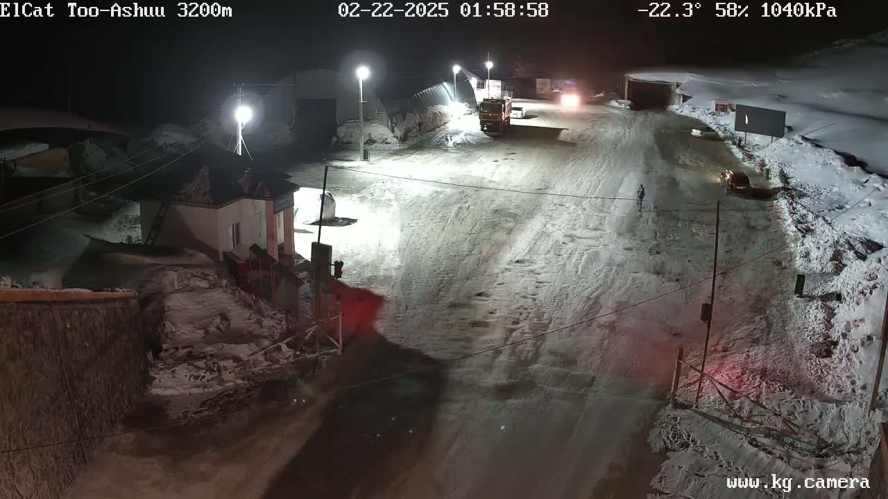 A snow-covered road at night, with a few vehicles and buildings visible under bright lights, at -22.3°C and 58% humidity.