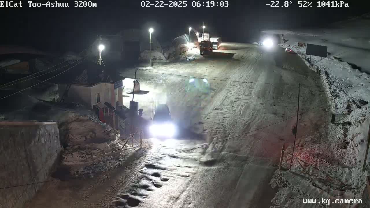 A snow-covered road at night is illuminated by streetlights, with a few vehicles visible, and a small building to the side.