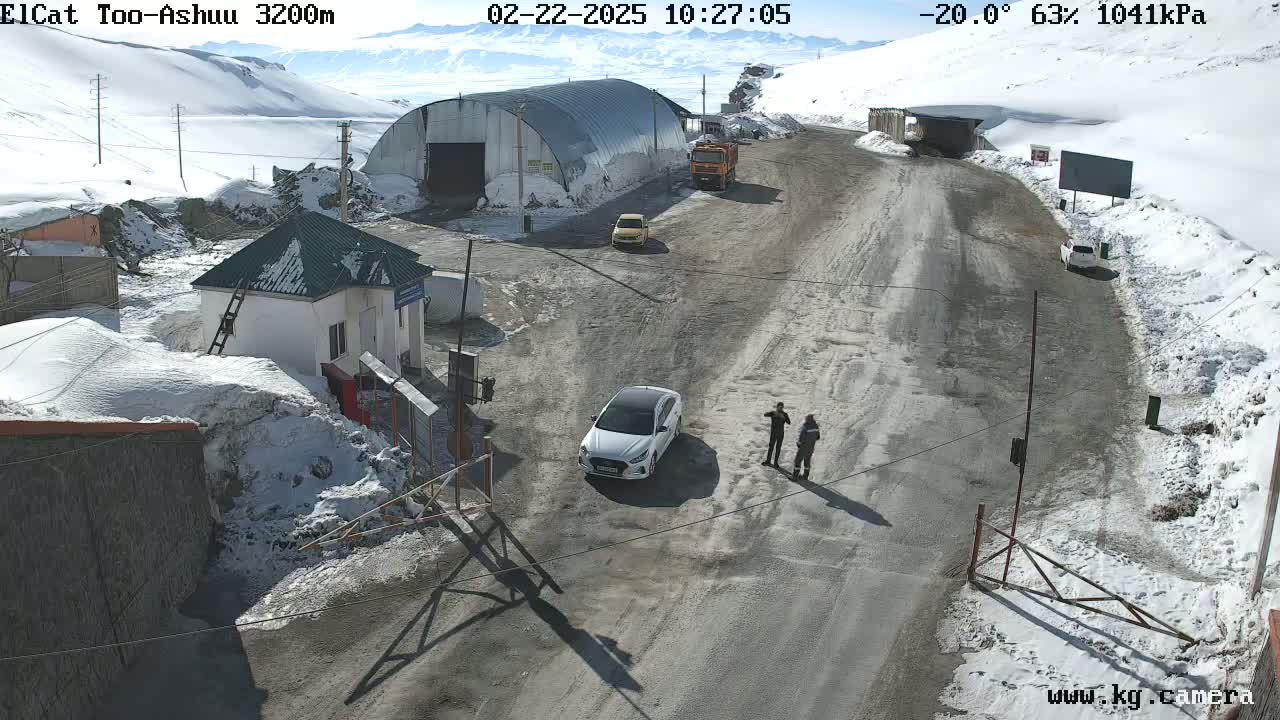A snowy mountain road passes by a small building and a large metal structure, with several vehicles parked nearby and two people standing near a white car under a sunny sky.