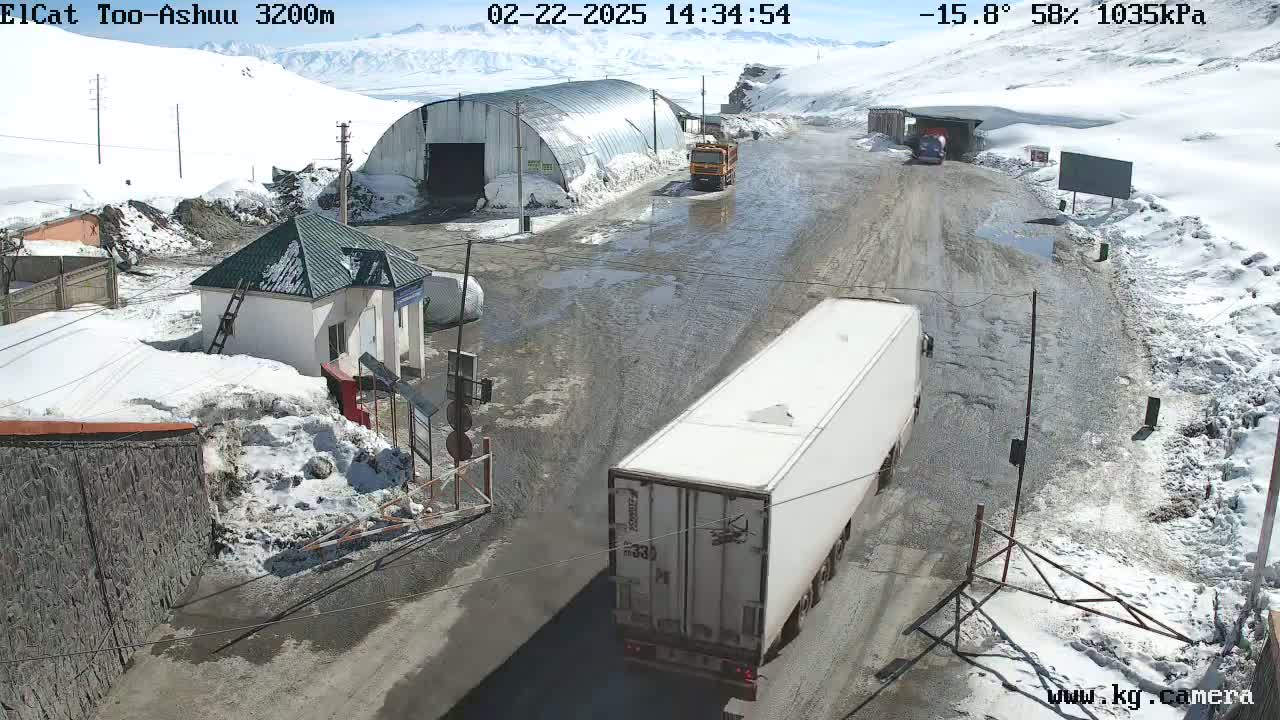 A snowy, mountainous landscape with a muddy road shows several vehicles near a small building and a large metal structure;  a white semi-truck is prominently featured in the foreground.