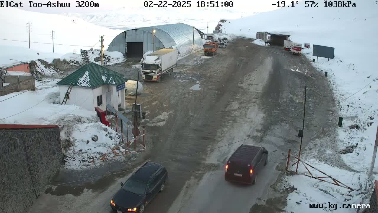 A snow-covered mountain pass with several vehicles near a checkpoint building and a large warehouse, under cloudy skies and a temperature of -19.1°C.
