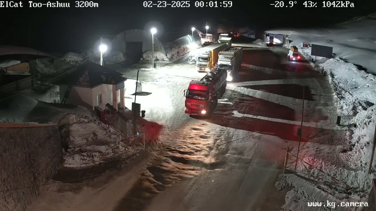 Several trucks are parked in a snowy, nighttime roadway near a small building.