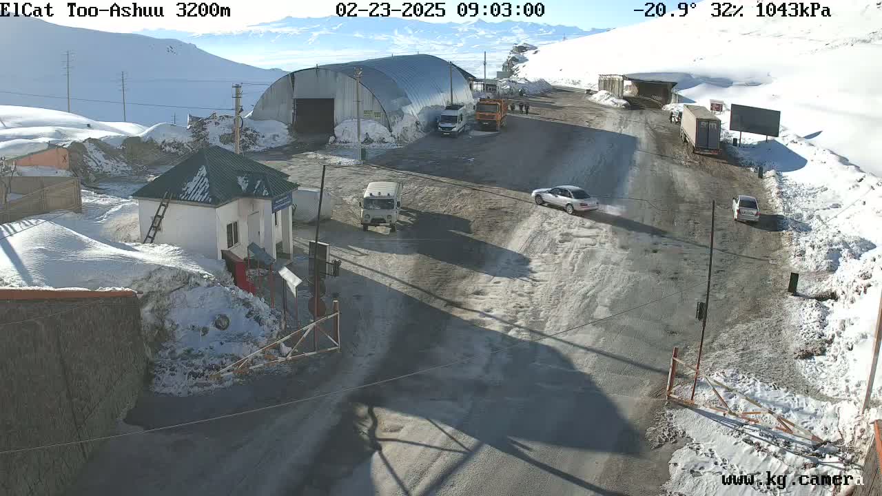 A snow-covered mountain pass road scene with several vehicles near a small building and a larger metal structure, under a sunny sky.